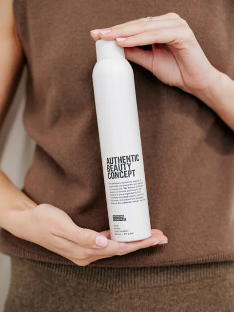 Person holding a white bottle labeled 'Authentic Beauty Concept' against a brown background